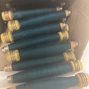 May include: A collection of teal-colored thread spools inside a cardboard box. The spools have wooden cores with yellow and silver metal accents. The thread is tightly wound around the cores, creating a cylindrical shape. The spools are arranged in a slightly haphazard manner.