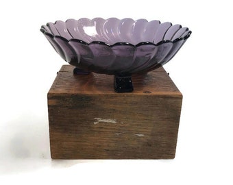 May include: A purple glass bowl with a scalloped edge and three feet. The bowl is sitting on a wooden surface.