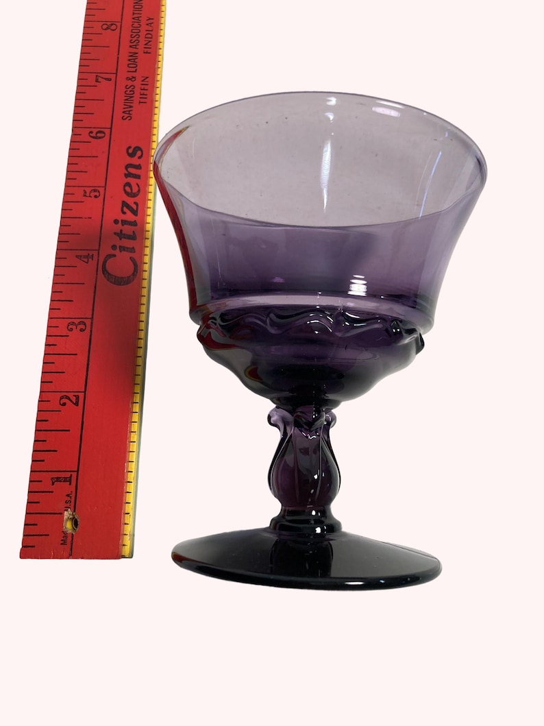 A purple glass wine glass with a flared bowl sits next to a red ruler on a white background.