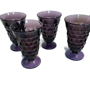 May include: A set of four purple glass goblets featuring a raised geometric honeycomb pattern on the exterior. Each glass has a wide circular rim, a tapered body, and a sturdy pedestal base, all crafted from translucent deep purple glass.
