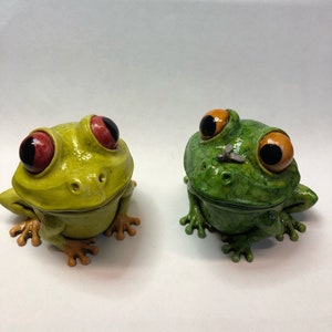 Può includere: Due statuine di rana in ceramica con grandi occhi espressivi. Una rana è verde con una mosca sul naso, l'altra è gialla.