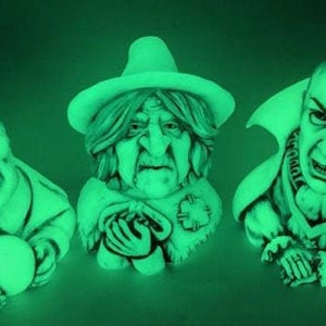 May include: Three green-toned Halloween character busts: Frankenstein, a witch, and Dracula. Frankenstein holds a round object. The witch wears a hat and holds a small creature. Dracula has a stern expression and a high collar.