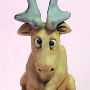 以下が含まれることがあります： 大きな表情豊かな目をした、小さなセラミック製のヘラジカの置物。ヘラジカは白い台座に座っており、茶色の毛皮と灰色の角をしています。