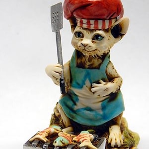 May include: A whimsical figurine of a cat chef wearing a red chef's hat and blue apron with a white star. The cat is grilling fish on a small barbecue. A salt shaker with "CAT" printed on it sits nearby.