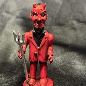 Może przedstawiać: Figurkę diabła w kolorze czerwonym z rogami, czarnym garniturem i srebrnym widłami. Figurka stoi na czerwonej podstawie.