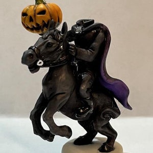 May include: A black horse figurine with a rider wearing a black cape and hat, holding a carved orange pumpkin. The figurine is on a white base.