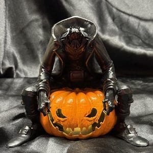 May include: A black figurine of a person wearing a long coat and hat, sitting on top of an orange pumpkin with a carved face. The pumpkin has a light inside, illuminating the carved face.