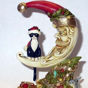 May include: A ceramic figurine of a crescent moon with a Santa face, a black and white cat wearing a Santa hat sitting on the moon, and a small gray rabbit sitting on a Christmas tree with presents at the base. The moon is decorated with gold paint and the tree is decorated with green and gold ribbon.