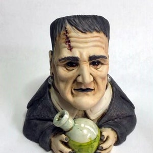 May include: A ceramic figurine of Frankenstein holding a green glass bong. The figurine is wearing a black suit jacket and has a stitched scar on his forehead.