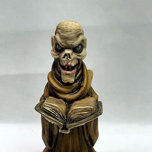 May include: A detailed figurine of a skeletal figure in a brown robe, holding an open book. The figure has blue eyes and a sinister grin. The base is white, and the book has gold-colored pages.