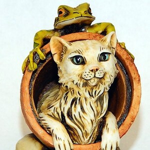 Può includere: Una statuina di gatto bianco che spunta da un vaso di terracotta, con una statuina di rana verde in cima. Una piccola statuina di topo si trova alla base del vaso, circondata da pietre ed erba verde.