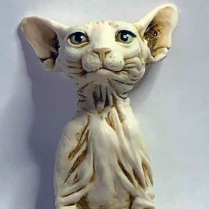May include: A white ceramic figurine of a hairless cat sitting with its ears perked up. The cat has a wrinkled face and large green eyes.