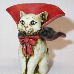 Peut inclure: Figurine de chat en céramique crème, déguisé en vampire. Il a les yeux verts, un nœud papillon rouge et une cape rouge. La figurine a des crocs et est en position assise.