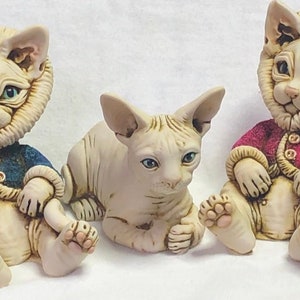 Pode incluir: Cinco estatuetas de cerâmica de gatos, todas com corpos sem pelos e roupas de diferentes cores. Os gatos estão todos em posição sentada ou deitada.