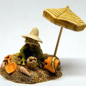 May include: A green frog wearing a brown hat sits on a sand dune with a yellow and white striped beach umbrella. The frog is holding a small orange bucket and shovel. There are seashells and a sandcastle on the dune.