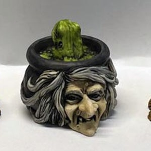 May include: Three ceramic cauldron candle holders featuring a witch's face, a frog, and a green plant. The cauldrons are black with orange flames and are displayed on a white surface.