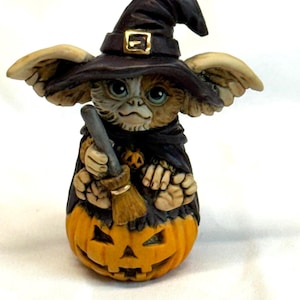 May include: A Halloween figurine of a creature wearing a witch's hat with a gold buckle and a black cloak. The creature holds a broom and sits on an orange jack-o'-lantern with carved features. A spooky decorative item.
