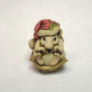 May include: A small, white ceramic figurine of a mouse wearing a red Santa hat with green holly and sitting in a brown, spiky chestnut shell.