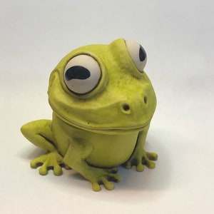 Può includere: Una statuetta di rana in ceramica verde con grandi occhi bianchi e un ampio sorriso con i denti. La rana è seduta con le gambe ripiegate sotto il corpo.