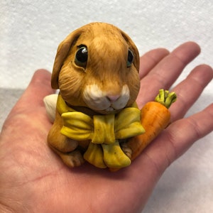 Può includere: Una piccola statuina di coniglio in ceramica marrone con un fiocco giallo e che tiene una carota.