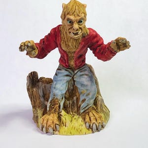 May include: A detailed figurine of a werewolf in a red jacket and blue jeans, posed on a tree stump. The werewolf has a snarling expression, with yellow fur and claws. The base is painted with green grass.