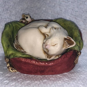 May include: A white cat figurine curled up asleep in a red and green bed with a small white mouse figurine beside it.