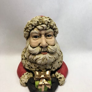 May include: A ceramic figurine of Santa Claus holding a green gift box with a gold bow. He is wearing a red suit with white fur trim and a white beard.