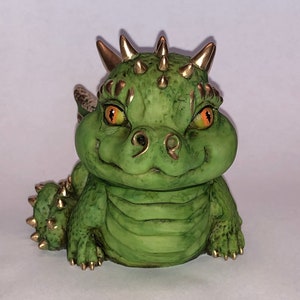 Può includere: Una piccola statuetta di drago in ceramica verde con dettagli dorati. Il drago ha un ampio sorriso e grandi occhi arancioni.