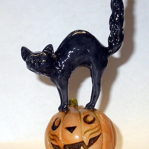 May include: A black ceramic cat figurine standing on top of a ceramic pumpkin with a carved jack-o'-lantern face. The cat is arched with its tail curled up.