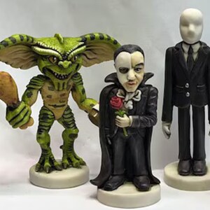 May include: A set of four hand-painted figurines featuring iconic horror movie characters. The figurines are painted in realistic detail and include a green and brown Gremlin holding a drumstick, a black-clad Phantom of the Opera holding a red rose, and a slender, white-faced Slender Man in a black suit.