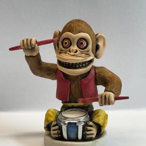 May include: A brown monkey figurine with red eyes and a toothy grin, wearing a red vest and yellow pants, plays a drum with two red drumsticks.