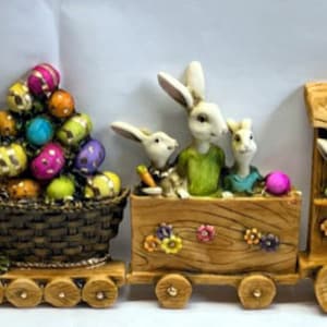 May include: A wooden toy train with bunny figures and a basket of colorful Easter eggs. The train has a brown wooden engine, a green and brown passenger car, and a brown wooden caboose. The bunnies are white with pink noses and are wearing colorful clothes.