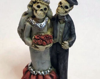 May include: A skeletal bride and groom wedding figurine. The bride is wearing a white dress with a red ruffled skirt and a floral bouquet. The groom is wearing a black suit and a top hat.