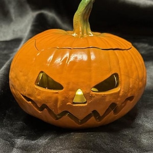May include: Orange ceramic pumpkin with a carved jack-o'-lantern face. The pumpkin has a green stem and a lit candle inside.