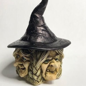 May include: A ceramic figurine of a three-faced witch wearing a black pointed hat with runes and symbols. The witch has long white hair and a green face.