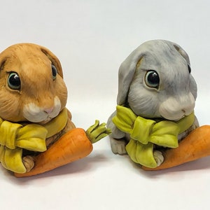 Può includere: Due statuine di coniglio in ceramica, una marrone e una grigia, entrambe con sciarpe gialle e che tengono carote arancioni.