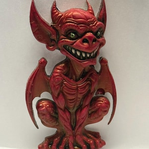 May include: A red gargoyle figurine with a menacing grin and green eyes. The figurine has bat-like wings and is in a crouching position. The sculpture is detailed with textured skin and horns.
