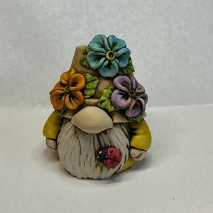 May include: A small ceramic gnome figurine with a flower pot hat. The hat is decorated with colorful flowers and the gnome has a white beard and a ladybug on its chest.