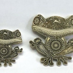 May include: Two intricately designed frog pins, one larger than the other, with a white background and black detailed patterns. The frogs are in a sitting position with their legs folded.
