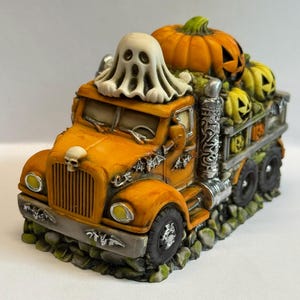 May include: A decorative orange truck figurine featuring a white ghost on the roof, a skull on the hood, and a truck bed filled with orange and yellow jack-o'-lantern pumpkins. The truck is adorned with silver bat accents and rests on a rocky base.