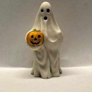 May include: A white ceramic ghost figurine holding a small orange pumpkin with a carved jack-o'-lantern face. The ghost has two black eyes and a wide open mouth.