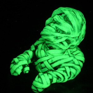Op de afbeelding: Een groene, in het donker lichtgevende mummiefiguur met een gedetailleerd, gestructureerd oppervlak. De mummie is gewikkeld in verband en heeft een licht geopende mond.