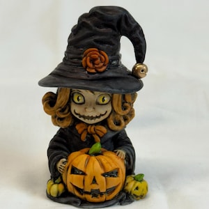 May include: A Halloween figurine of a witch with curly hair, wearing a black hat with a rose and a bell. She holds an orange jack-o'-lantern with a carved face, and is flanked by two smaller pumpkins. The witch is dressed in a black outfit.