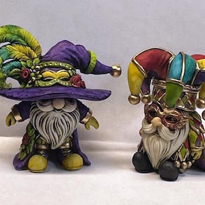 May include: Two Mardi Gras gnomes, one wearing a purple hat with green and yellow feathers and a mask, the other wearing a multi-coloured jester hat with a mask.