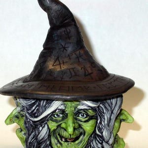 May include: A ceramic witch figurine with a pointed hat covered in runes and symbols. The witch has a green face, white hair, and a mischievous grin. She is holding a small green creature in each hand.
