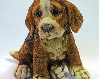 May include: A brown and white ceramic figurine of a sad-looking beagle puppy sitting with its paws crossed. The puppy has a white patch on its forehead and brown fur.