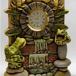 Puede incluir: Un reloj de cerámica con una cascada y dos ranas. La esfera del reloj está rodeada por un diseño de piedra con nenúfares y flores. La esfera del reloj dice "QUARTZ".