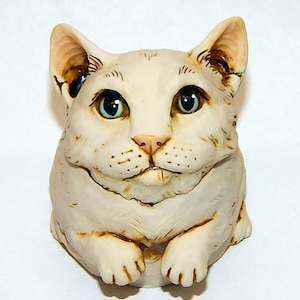 Può includere: Statuetta di gatto color crema con dettagli. Il gatto ha grandi occhi verdi espressivi, un naso rosa e dettagliate marcature del pelo. La statuetta è in posizione seduta, con le zampe visibili.
