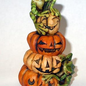 May include: A vertical stack of four orange carved pumpkins with carved facial expressions. A green frog with dark stripes is perched on top of the highest pumpkin, and a second green frog is positioned on the side of the bottom pumpkin.