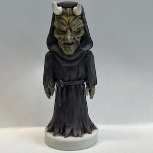 May include: A green and black figurine of a demon wearing a robe with a white base. The demon has horns and a menacing expression.